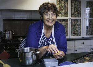 I am a woman in my late sixties with fair skin and short dark hair, in my kitchen leaning on my countertop smiling warmly at the camera while preparing dough with my hands – I am baking cookies. I am wearing an electric blue long sleeve top and a stripey navy and white apron. To my right there is a metal pot on the dark green countertop. To my left, a baking tray with cookie dough portions sits ready for the oven. Behind me, the traditional kitchen is decorated with floral-patterned curtains inside glass-front cabinets and a stove with shiny pots. The overall mood suggests warm, welcoming homely charm - it feels like a snapshot of me happily sharing my cooking.