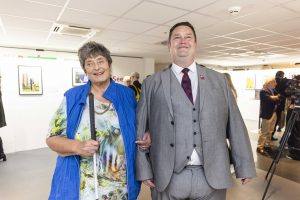 Penny is with Clive Jones. Their arms are linked and they are both smiling to camera, stood in the middle of the exhibition